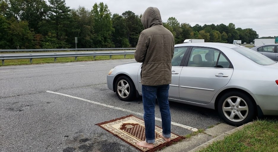 A person preparing for prayer on a travel mat near a car on a highway, illustrating the need for portable wudu solutions.